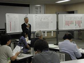 1人の男性が3つに並べられたホワイトボードの前でマイクを持ち話をしている所を他の参加者の人達が聞いている写真