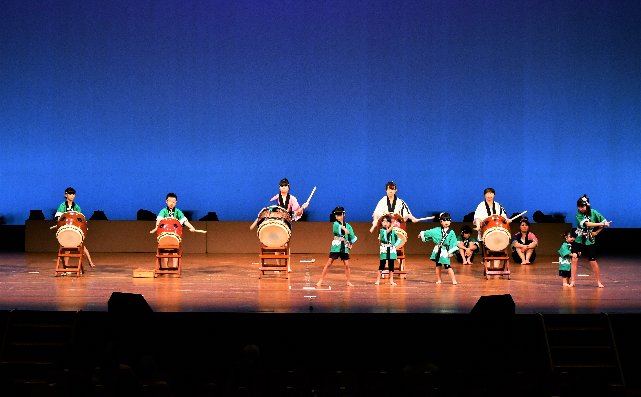ステージ上で、法被を着た大人や子供が、太鼓をたたいていたり、踊りを披露している様子の写真