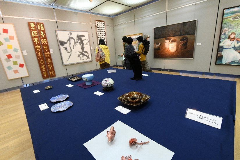紺色のクロスが敷かれたテーブルの上に陶器が展示され、壁に絵画や書道などが展示されている芸術展の写真