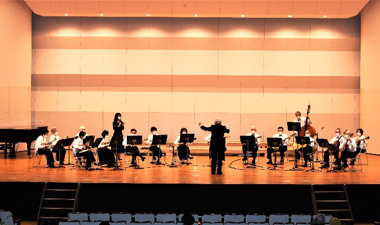 ステージ上で指揮者が指揮を振り、コントラバスや、弦楽器などで演奏が行われている写真