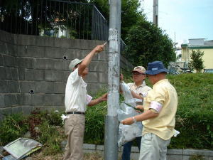 1名の男性がごみ袋を持ち、2名の男性がスクレーパーのような道具を使い電柱に貼られた違法屋外広告物を除去している写真