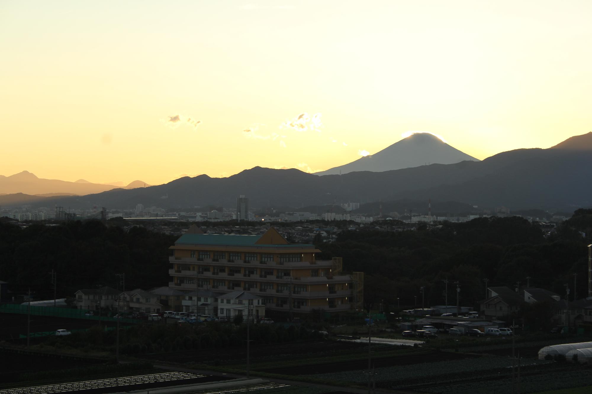 住宅や田んぼや畑、辺りは薄暗く太陽の明かりで黄色く染まった空と山々の向こうに見える富士山の写真