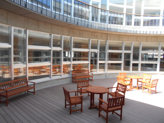 窓ガラス側に2脚のベンチ、手前に4脚ずつ椅子がセットされた2台の丸テーブルが設置されている1階中庭の写真