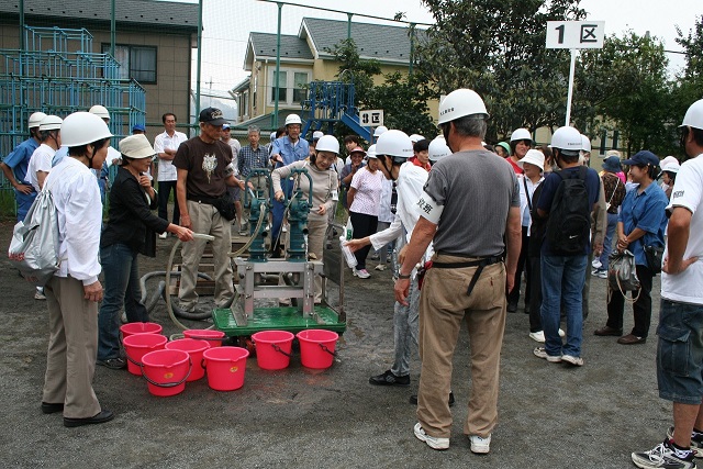 ポンプでバケツに水を汲んでいる女性やヘルメットを被って集まっている防災訓練の参加者の写真