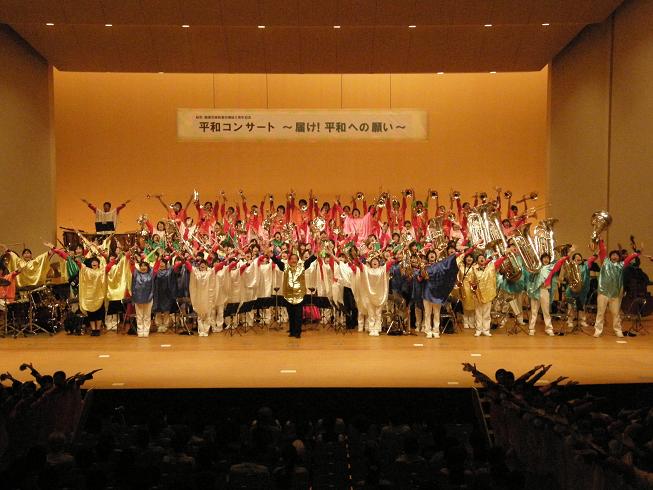 黄色、青、白、緑、ピンクとカラフルな衣装を着た市立柏高等学校吹奏楽部の人達が舞台上で手を挙げ挨拶をしている写真