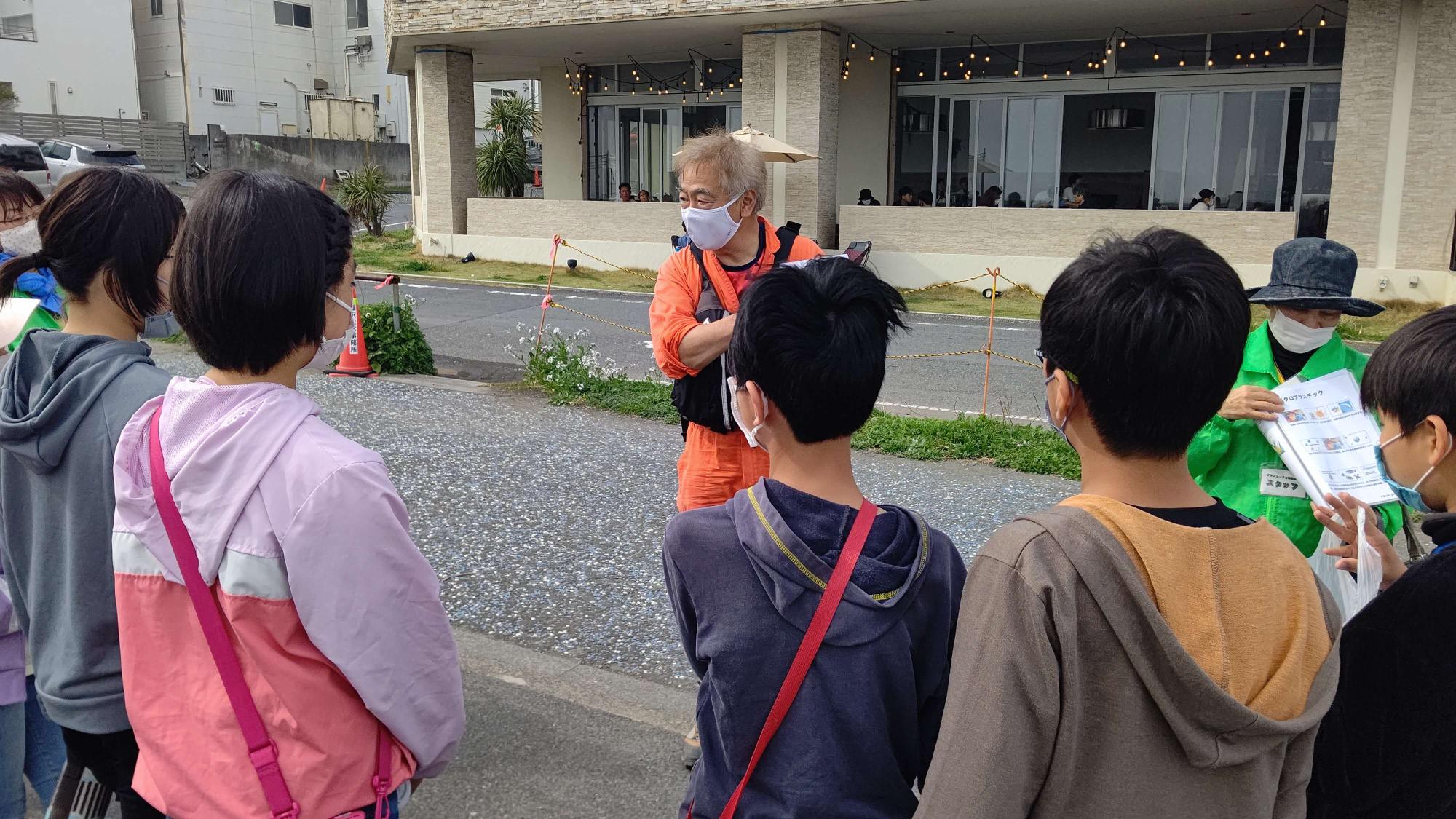 参加した子供たちがオレンジ色のつなぎを着たおもしろ博士の話を聞いている様子の写真