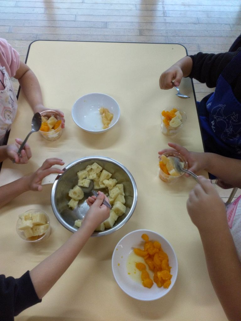 4人の園児がテーブルに座り、角切りにしたスポンジケーキやフルーツを自分のカップにお好みで盛り付けている写真