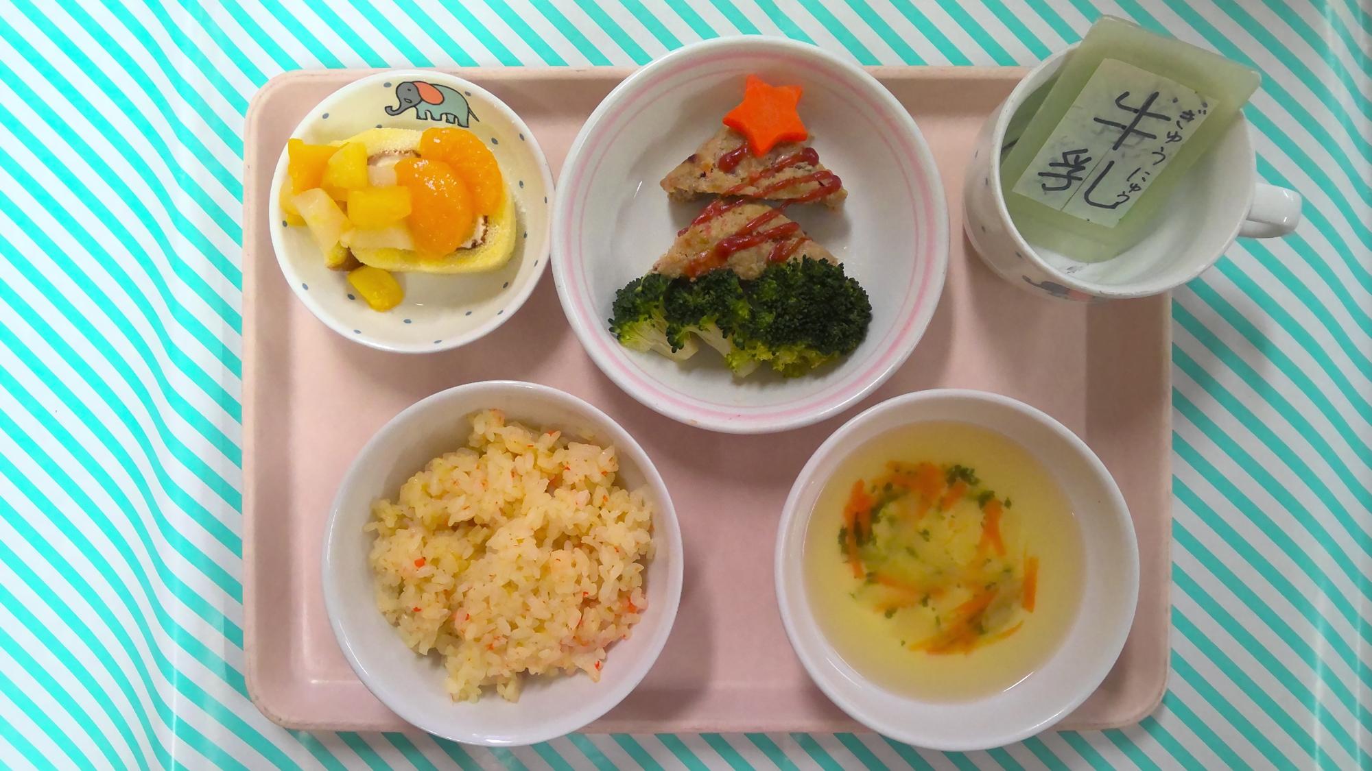 左側お茶碗にキャロットライス、右側お椀に野菜スープ、左奥にロールケーキの上に角切りにしたフルーツ缶をのせたクリスマスケーキ、中央奥のお皿にツリーに見立てたミートローフにケチャップで模様を描きツリーのトップに星形にんじんをのせ、根元の部分はゆでたブロッコリー、右奥、牛乳と書いた札が入ったコップがトレーに並んでいる写真