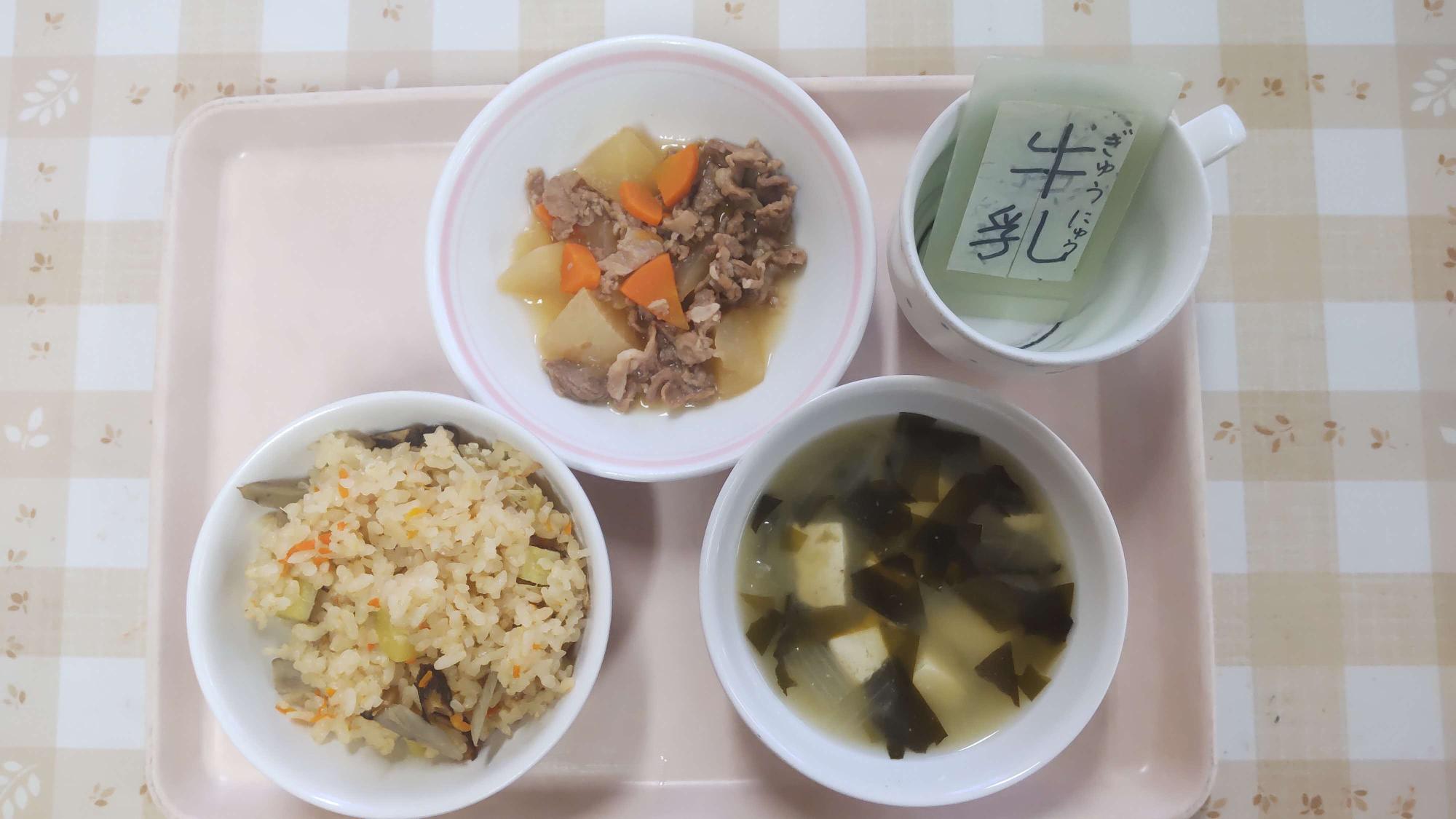 左茶碗に野菜を炊き込んだ吹き寄せごはん、右側のにわかめと豆腐のみそ汁、奥中央に豚肉と大根の炒め煮、奥右側に牛乳と書いた札が入っているコップがトレーにのっている写真