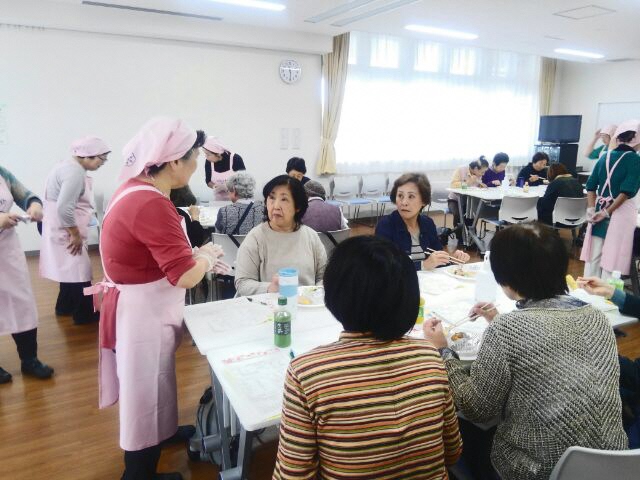 テーブルごとに座っている試食会の参加者が、ピンク色のエプロンに三角巾をつけた女性スタッフに説明を受けている写真