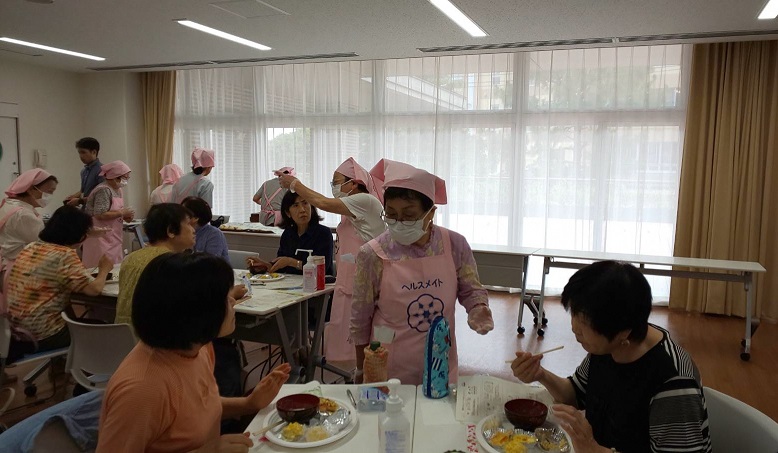 試食会にて食事をしている人や、ピンクのエプロンと三角巾を着けた方と話をしている試食会参加者たちの写真