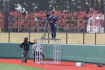 野球場の外野スタンド側のフェンス前に男性の背丈ほどの足場を組み、その上にカメラを設置し男性が立っている写真