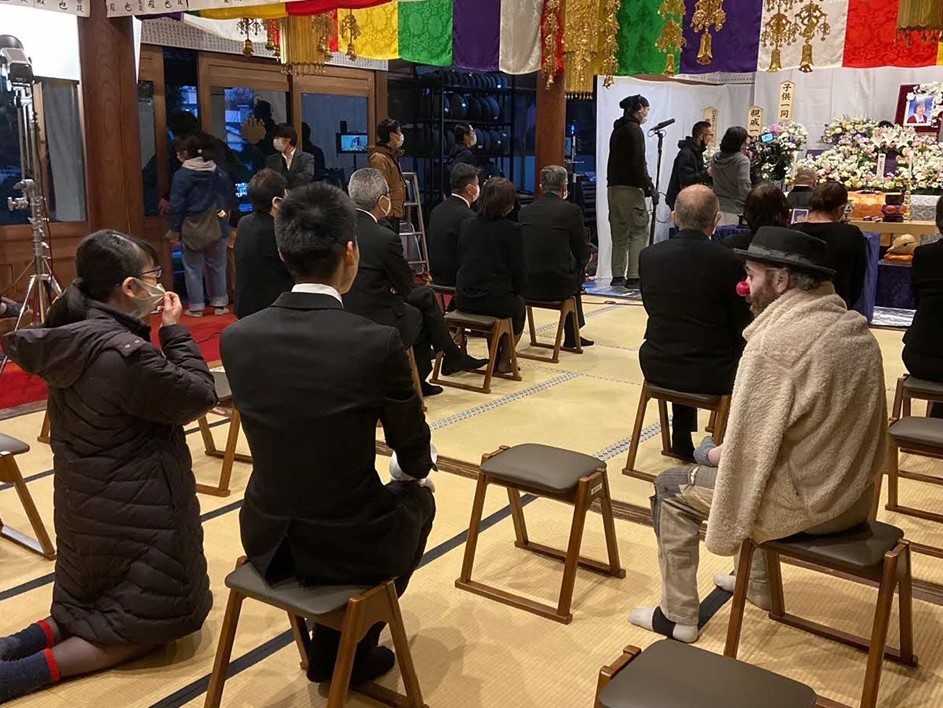 前方に祭壇、出演者の方々が和室に設置された椅子に座り、周りでスタッフの方々が準備をしている様子の撮影現場の写真