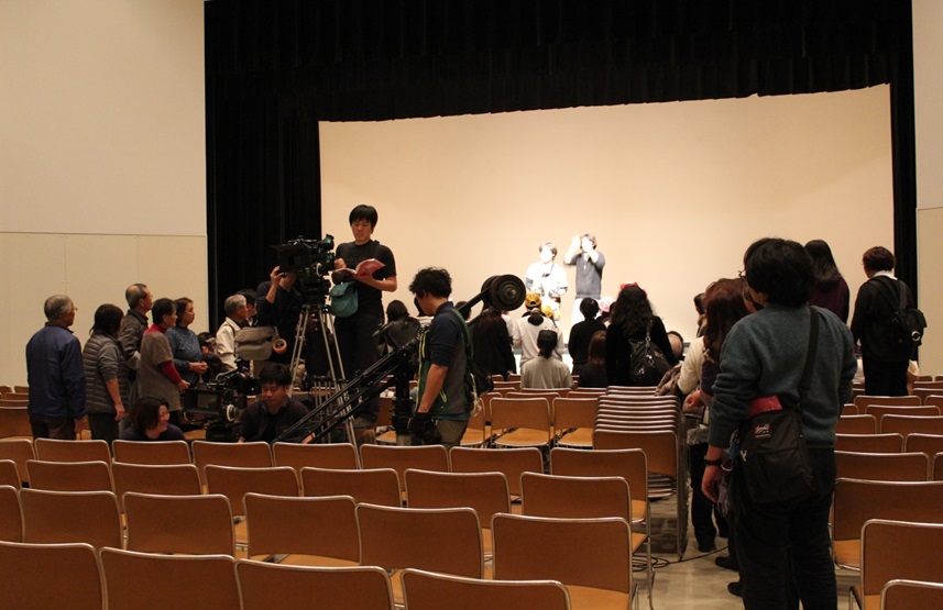 ホールの客席でカメラをセットしているスタッフや舞台のほうを向いて立っているエキストラの皆さんの写真