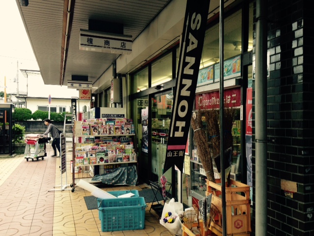雑誌や竹ぼうきが置かれているお店の入り口を店外入口右側から撮影した写真