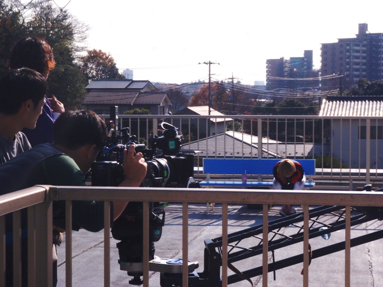 屋上のベンチに座っている俳優さんをカメラで撮影しているスタッフの写真