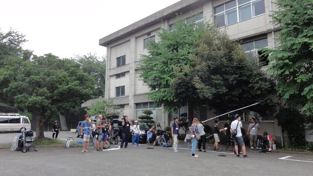 横に木が植えられている校舎の前に、カメラやマイクなどの記事を持ったスタッフの方々が集まり立っている写真