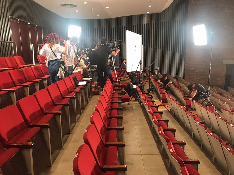 照明や、カメラなどがホール会場に持ち込まれ、赤い椅子に座っている人たちを撮影している写真
