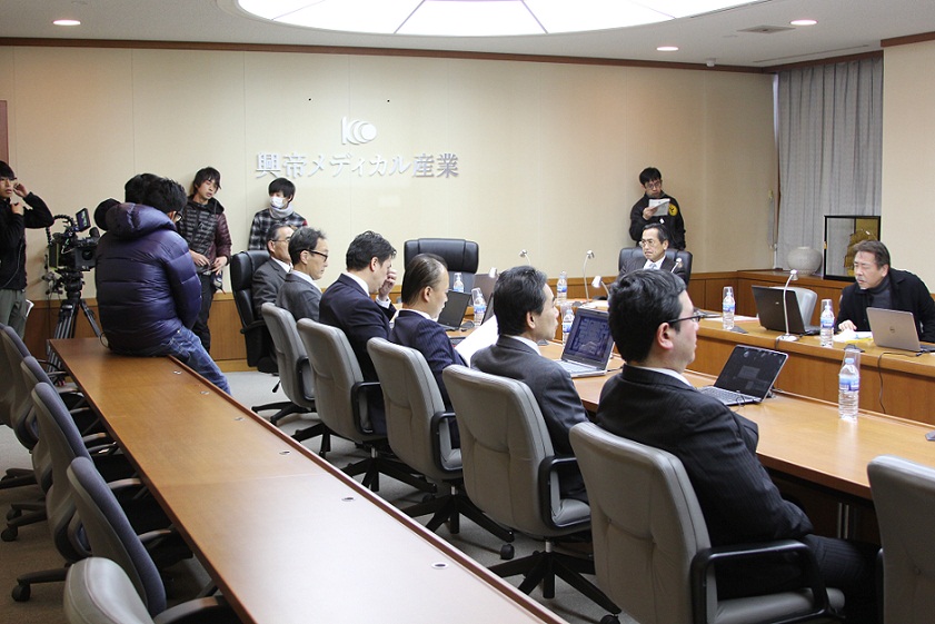 会議室にスーツ姿の6名の男性と反対側に2名の男性が座っている写真