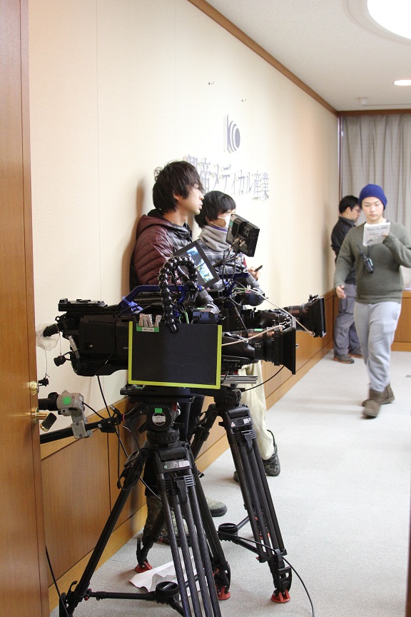 壁際に2名の男性が立っている手前に2台のカメラが置かれている写真