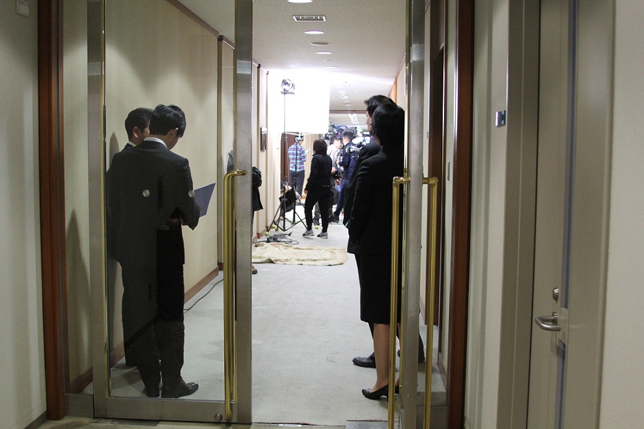 奥で照明が照らされ撮影が行われている手前のドアの近くでスーツ姿の4名の人が奥へ体を向けて立っている写真
