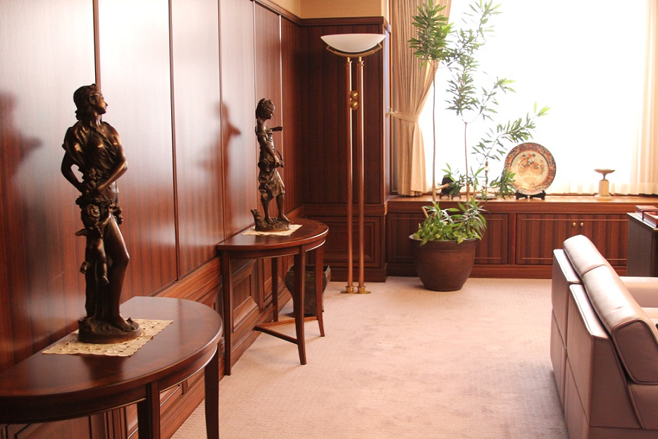 右側に革張りのソファ、奥の窓ガラスの手前に大皿や観葉植物、左側の壁際に2台の半円状の台の上にブロンズ像が飾られた社長室のセットの写真