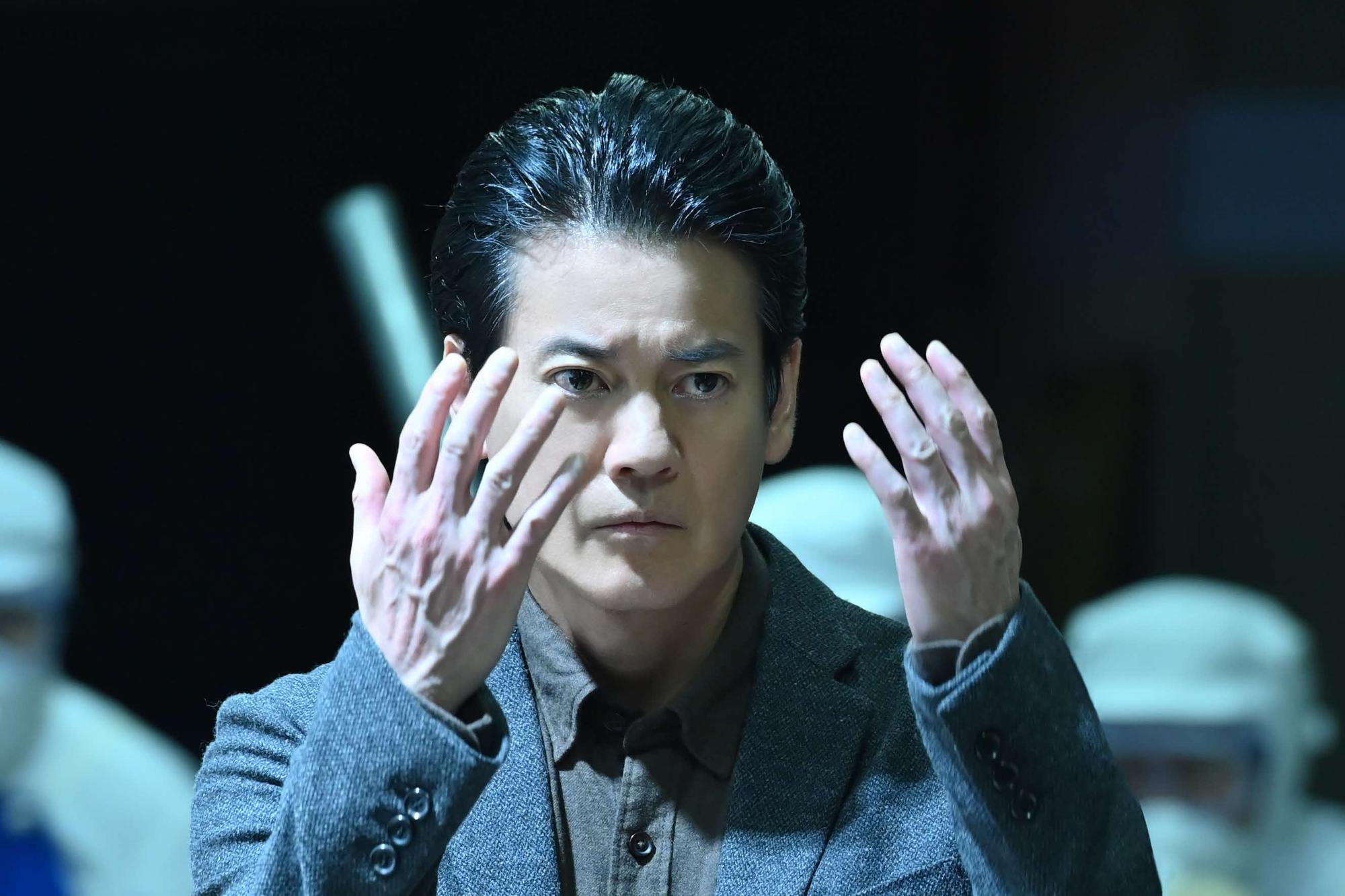 俳優 唐沢寿明さんが両手を顔の前にあげ、真剣な表情を見せているドラマの一場面の写真