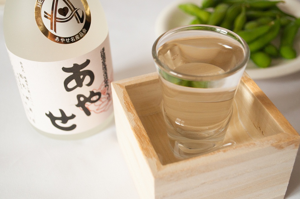 瓶にあやせのラベルがついた日本酒と、木桝の上に置かれたガラスコップに日本酒が注がれた写真