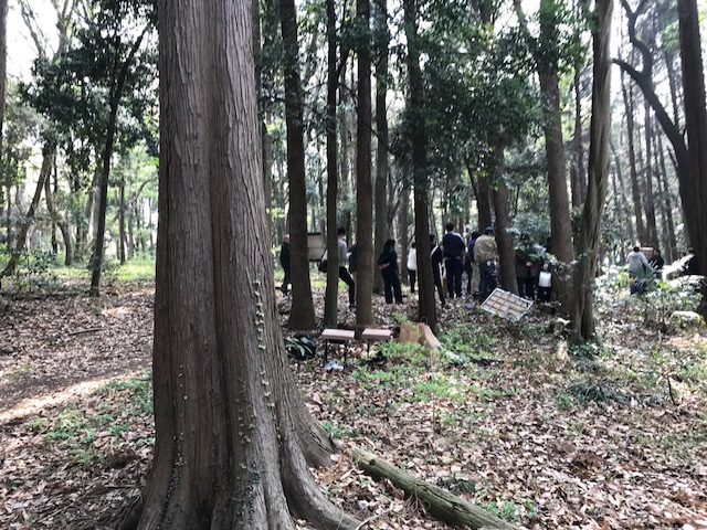 林の中で大勢のスタッフが撮影を行っている様子を1本の木の手前から撮影した写真