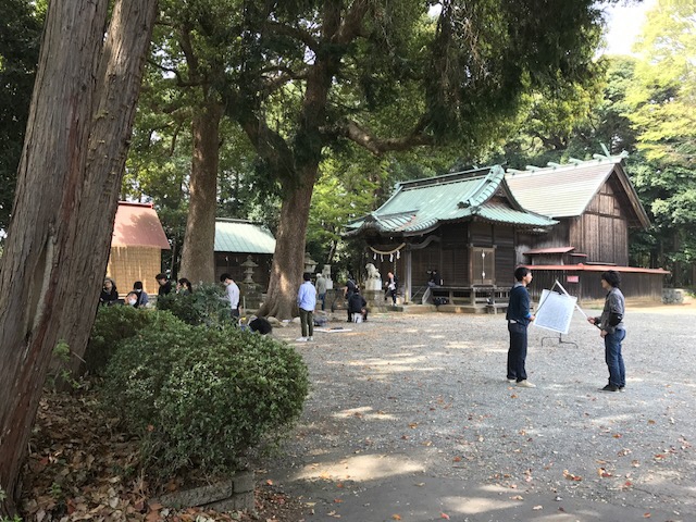大きな木に囲まれた神社の拝殿の前で撮影を行っているスタッフの人達、右側の広い境内で2名の男性が刀の先を合わせて練習を行っている様子の写真