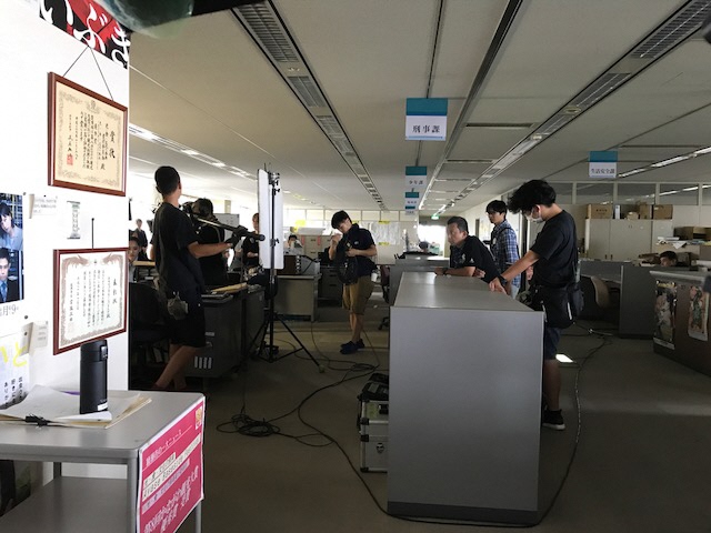 広いオフィスの一角で、マイクや配線などを持ったスタッフの方々が写っている撮影の様子の写真