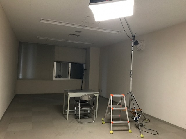 部屋の中央にグレーの机と向かい合わせて座るようにパイプ椅子が置かれ、その隣に脚立と大きな照明が置かれている取調室のような部屋の写真