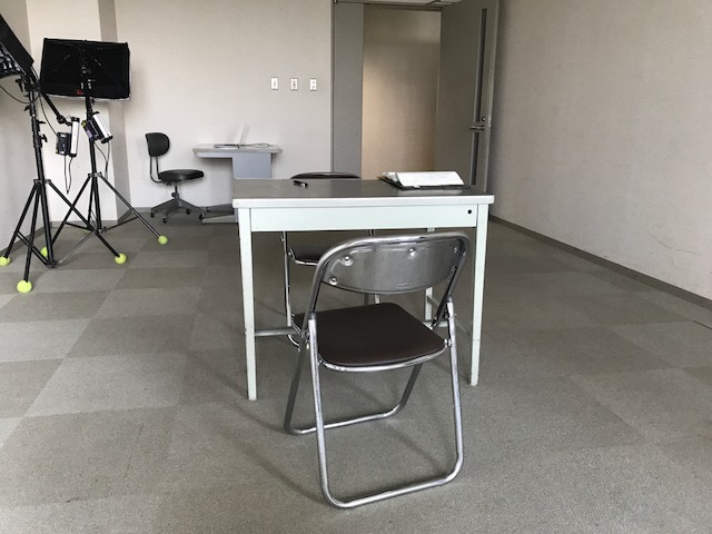 部屋の中央にグレーの机と向かい合わせて座るように置かれたパイプ椅子がある、取調室のような部屋の写真
