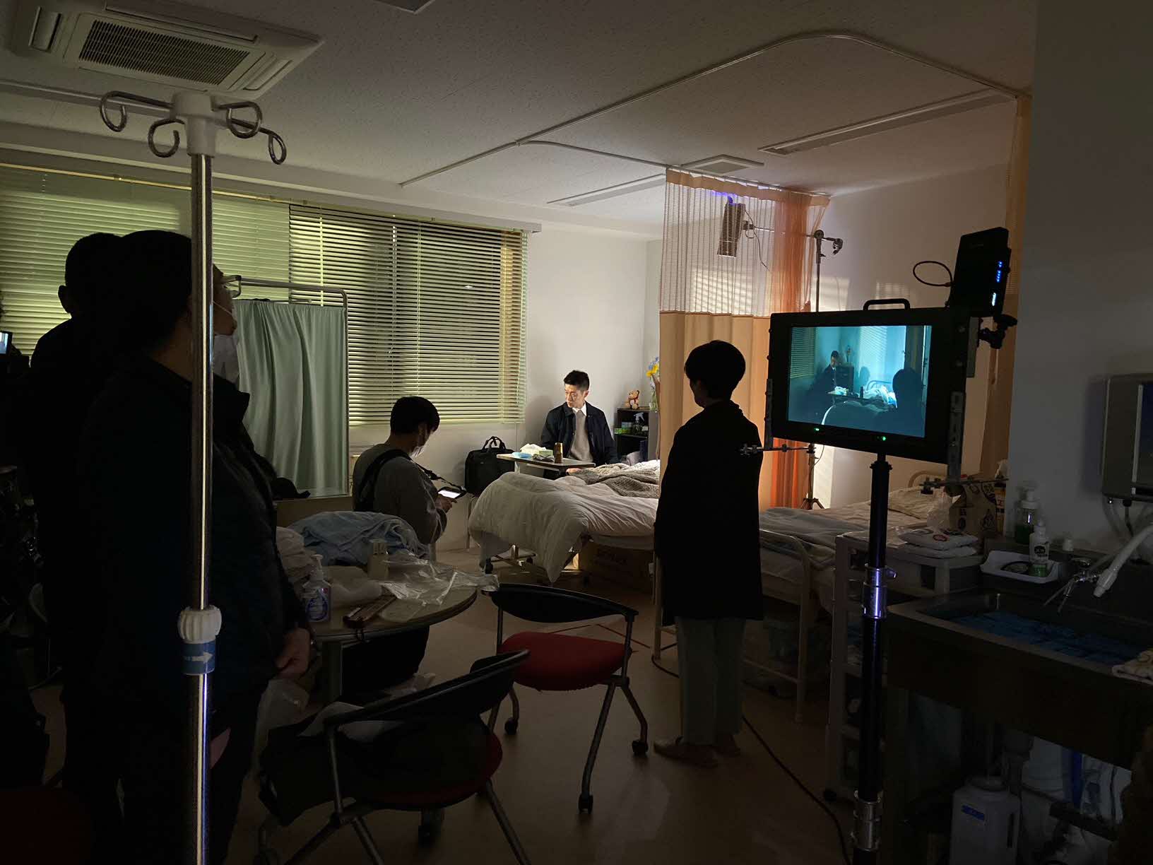 手前にモニターがあり、奥で役者の方がベッド横の椅子に座っている薄暗い病室での撮影現場の写真