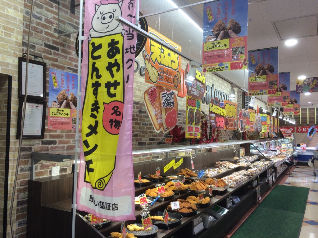 「あやせとんすきメンチ」と書かれたピンク色ののぼり旗の奥に揚げ物が並んでいるMEGAドン・キホーテ綾瀬店の店内の写真