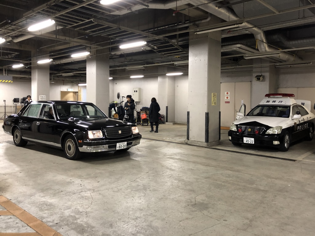 屋内駐車場の駐車スペースにパトカー、通路に黒いセダンの車が停まっている撮影現場の写真