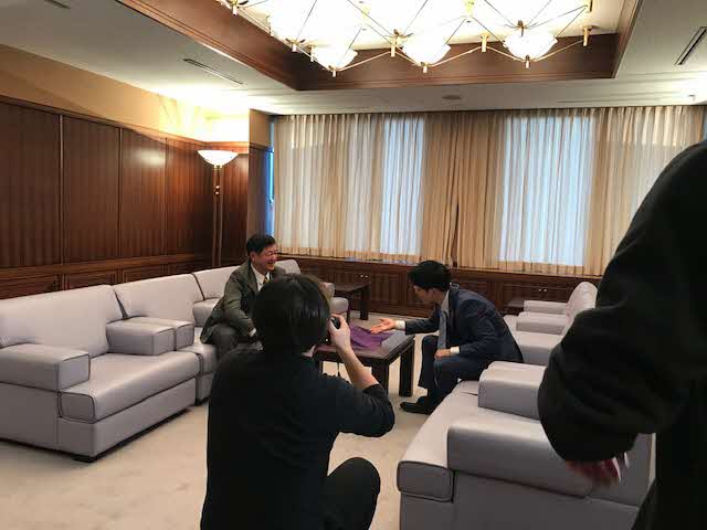 テーブルを挟んで2名の男性がソファに座っている様子の撮影が行われている写真