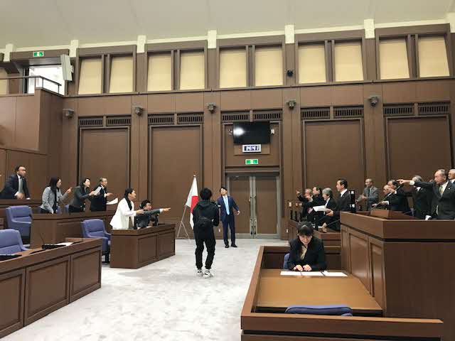 議会の場で黒色の服の男性にスーツ姿の男性が詰め寄り、左右の席の議員が立ち上がって指を指して言い争っている撮影の様子の写真
