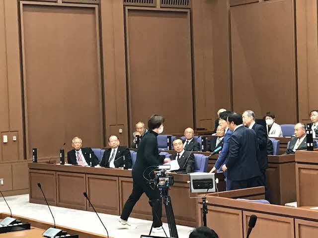 議員が座っている席に逃げたスーツ姿の男性に黒色の服を着た男性が詰め寄っている場面を撮影している様子の写真