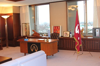 大きな窓の前に市長席がありその横には作品に登場する架空の自治体である八萬市の市旗が飾られた、市長の部屋を右斜めから写した写真