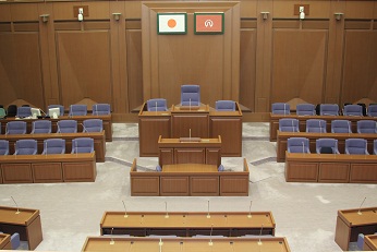 中央に日本国旗と作品に登場する架空の自治体である八萬市の市旗が掲げれた演台と議長席が中央にあり、両隣に議員席、視聴席が設けてある議会事務局内の写真