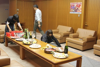 ソファーを壁際によせ、中央には長方形の机を2脚くっつけてテーブルの上に飲み物や食べ物の準備をしている3名スタッフの人の写真