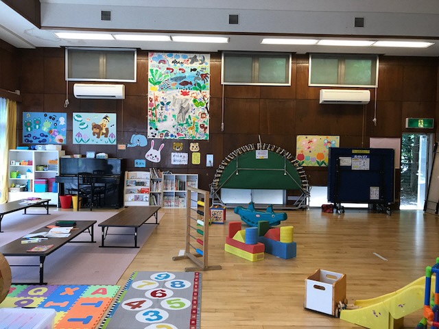 壁にはカエルや金太郎や魚などの絵が貼られ、子ども達の遊び道具や絵本が棚に収納されている、左側のスペースは絨毯の上に長机が置かれ、右側のスペースは自由に遊べるフローリングになっている、ながぐつ児童館の室内写真