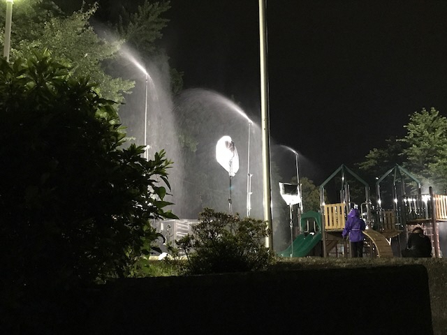 夜の公園で雨を降らせる機械を使って雨を降らせ、設置された照明機材で雨の降る夜の公園を照らしながら撮影を行っている様子の写真