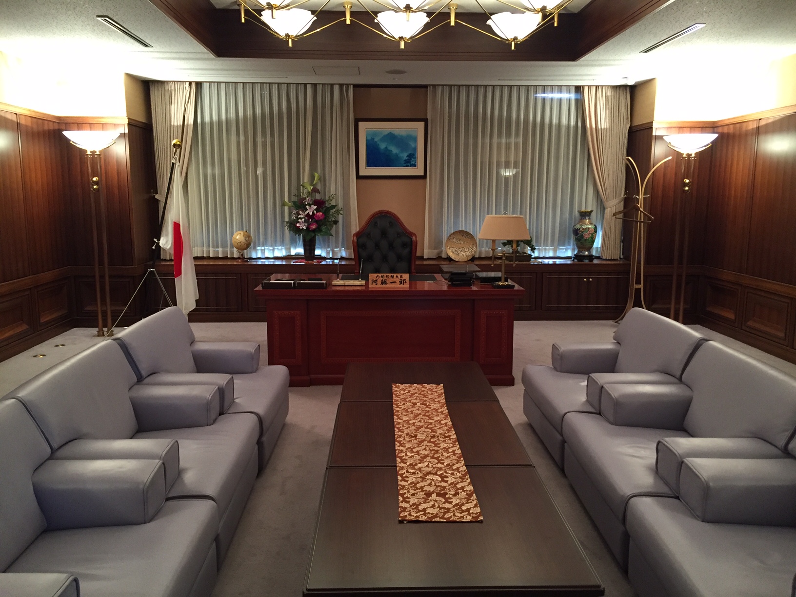 グレーのソファーが対面に並び、中央に同じ長さの長机、真正面に立派な机と左側に日の丸の旗が置かれた室内全体を写した写真