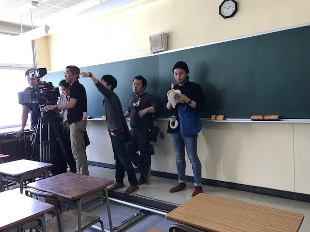 教室の黒板の前にカメラとスタッフの方々が集まり、撮影を行っている様子の写真