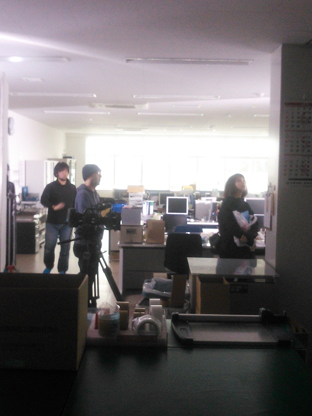 女性と男性がいる会社内を撮影をしているカメラマンを横から写した写真