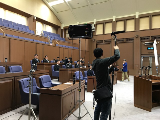 議場に撮影機材が設置され、議員席にエキストラの方々が座っている撮影風景の写真