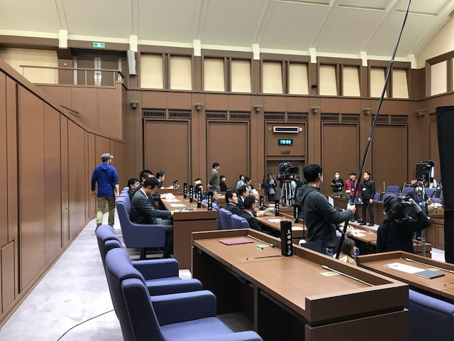 議員席に議員のエキストラの方々が座り、カメラや照明器具等の機材を持ったスタッフの方々が撮影の準備をしている様子の写真
