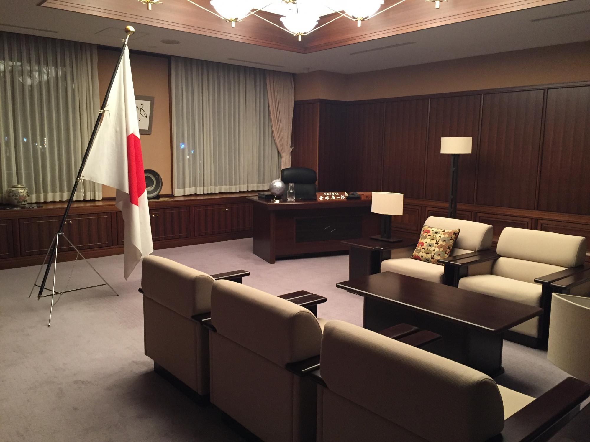 内閣総理大臣の机と椅子が室内奥にあり、左側に日の丸の国旗、右前方に応接セットのソファーと机が置かれている室内の写真